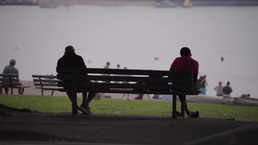 Two people sit on a bench watching the activity at the beach