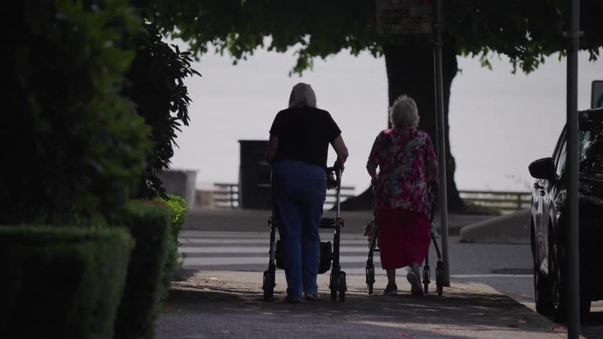 Two elderly women with rolling walkers slowly make their way down a shaded sidewalk towards a body of water