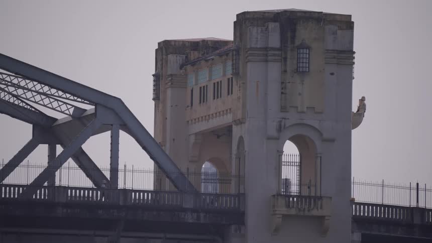 People and traffic on an old concrete bridge tower with arched openings and weathered walls stands beside a steel truss section of the bridge