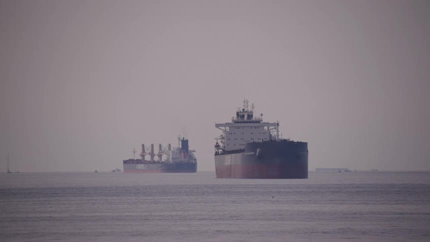 The video shows a wide shot of two large cargo ships sitting in the water on a hazy or foggy day the sky and water are muted gray and the shoreline is not visible