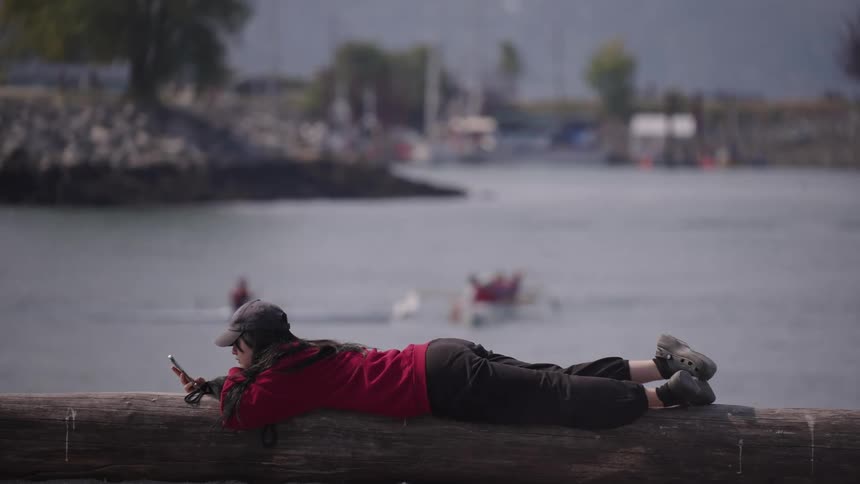 The video shows a person in a red shirt and black pants lying on a large log near the water they are holding a phone and looking at the screen with a blurry waterfront and some boats in the background