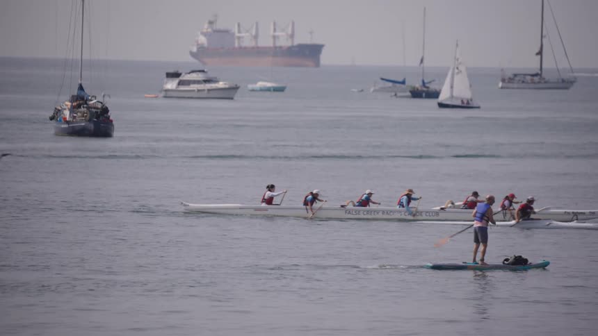 A man wearing a life vest is paddleboarding in the foreground while a team rows an outrigger canoe nearby and various sailboats and a large cargo ship are anchored in the background