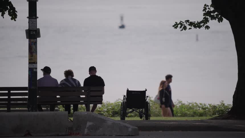 Three people sit on a bench on a sidewalk watching a lake in front of them while people walk past and cars drive along the road behind them with a wheelchair beside the bench at midday
