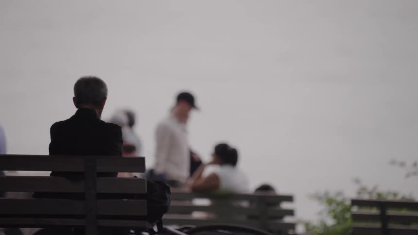 An older man sits alone on a bench facing the water while blurred figures in the background walk and talk nearby