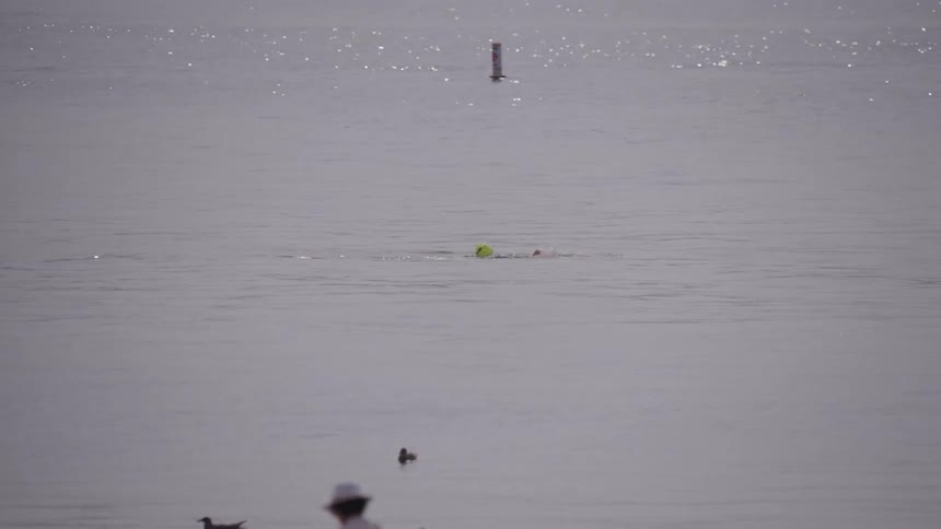 Person swimming in the sea with colorful cap on while others watch from the beach and a kayak floats in the background