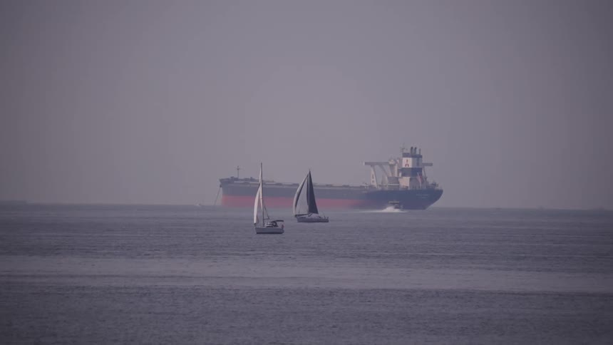 Two sailboats with sails up move on a body of water in front of a cargo ship in the background