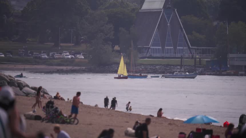 People enjoy the beach while a yellowsailed sailboat sails near the shore
