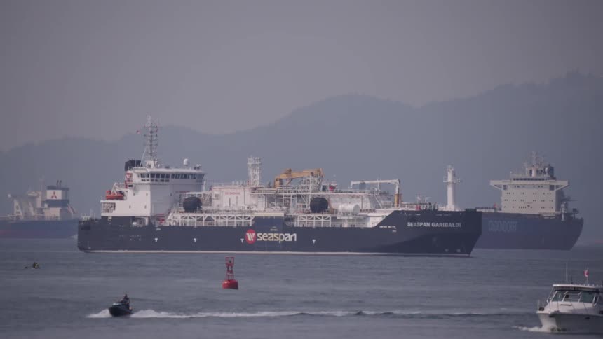 Jet skis and a motorboat travel through the water while freighters float near the shore on a foggy day