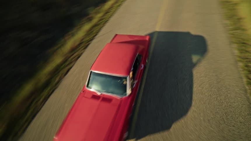 A car drives along a road that curves through a landscape of rolling hillsides with water and mountains in the background