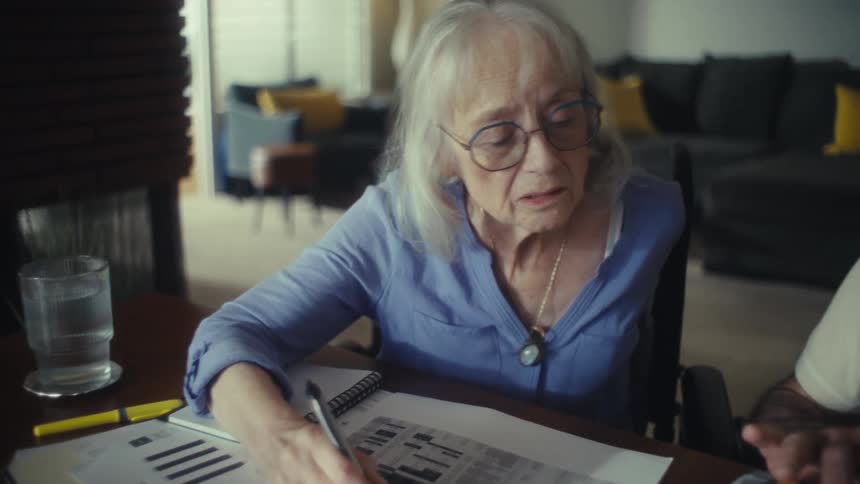 Elderly woman with glasses writes in a notebook while man beside her does calculations
