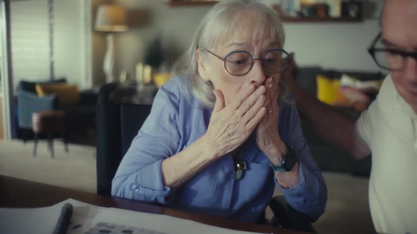 An older man and woman are sitting at a table with papers and a calculator the woman who has gray hair is looking at the papers and holding a pen the man also with gray hair is looking at her with a surprised expression hes wearing a creamcolored