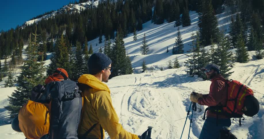 Friends stand near the base of a snow covered forested mountain wearing backpacks and having a conversation