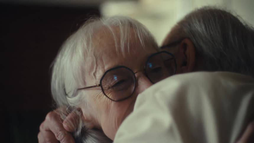 An older couple with gray hair and glasses is smiling at each other the woman is wearing a blue shirt and the man is wearing a creamcolored shirt they are sitting close together they hug warmly and continue to smile as they talk