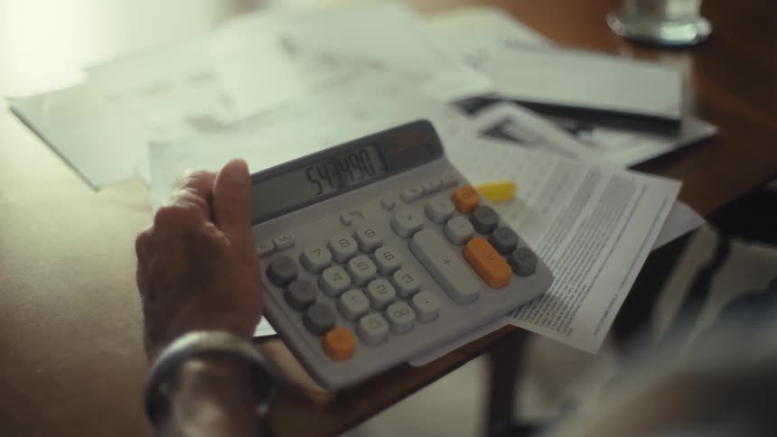 A persons hand holds and operates a calculator on a table with a stack of papers
