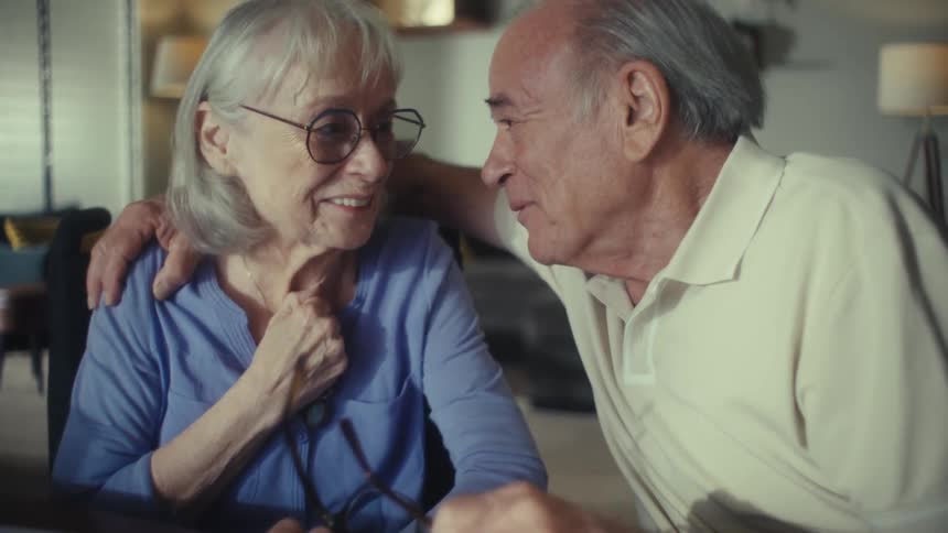 A couple with gray hair is seated at a table with the mans arm around the womans shoulder as they both smile and laugh while looking down and talking