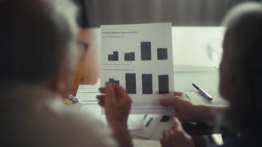 Two elderly people sit together reviewing a financial document with bar graphs comparing returns and risks