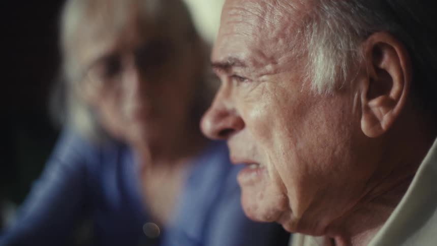 An older man and woman are sitting together the man is looking to the side with a pensive expression and the woman is looking at him with a look of concern the man has gray hair and is wearing a creamcolored shirt while the woman has gray hair and is
