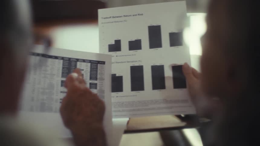 The video shows two people seated at a table with one person holding papers with charts and graphs on them they appear to be looking at and discussing the documents