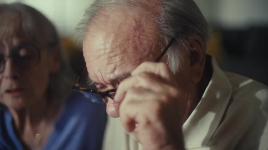 The provided video is only 4 seconds long and depicts an older man and woman both looking concerned the man who is closer to the camera appears distressed and at the 2second mark he puts on his glasses the woman in the background is blurry but als