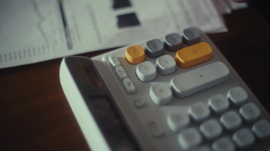A persons hands highlight a chart on a document and then press buttons on a calculator sitting on a table next to the papers