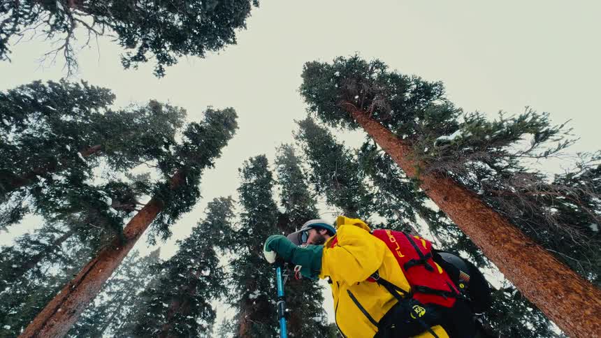 A young man walks up a snowy hill at midday wearing ski boots a helmet goggles a hat and a yellow jacket while holding ski poles surrounded by trees