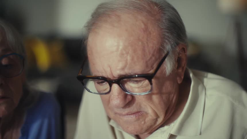 A senior couple appears worried and stressed while reviewing documents together culminating in the man taking off his glasses with a look of concern