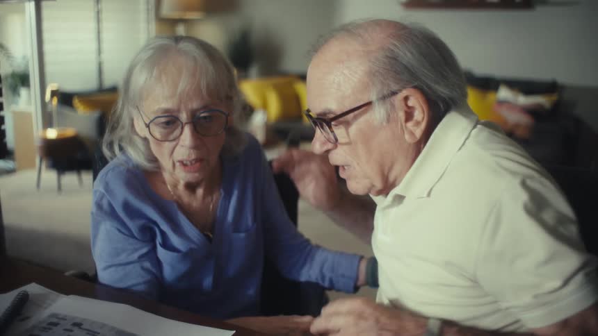 The video shows an older couple sitting close together on a couch they are both looking in the same direction appearing to be surprised or concerned by something out of frame