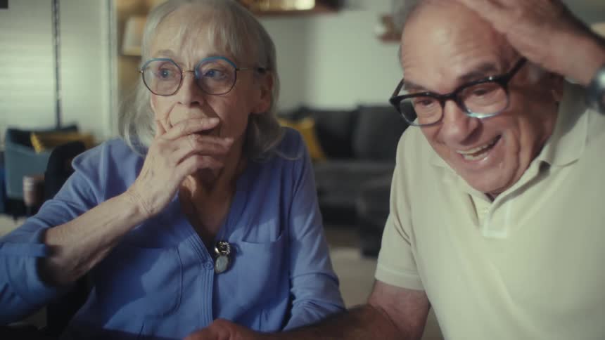A man and woman sit at a table and look at a calculator as the man taps a number and they both look up at each other with expressions of surprise and then happiness