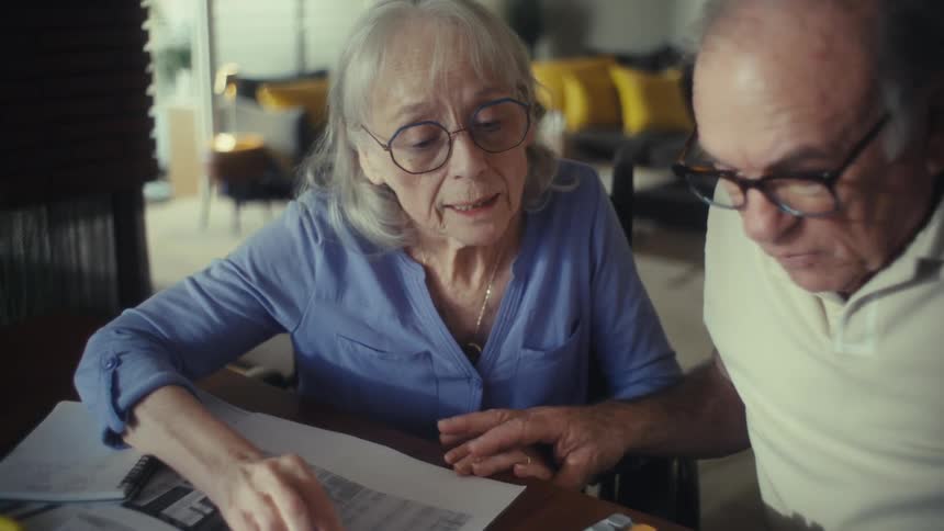 An elderly couple both wearing glasses sits behind a desk going through documents the man punches numbers into a calculator and the result excites them making them hug and smile at each other at midday