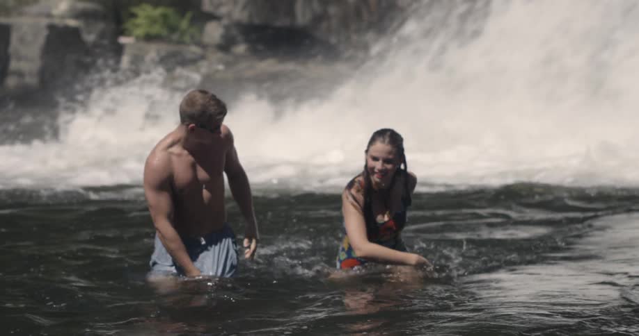 A man and woman wade in the water by a waterfall