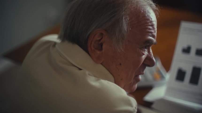 A man sits at a table and looks at a document with a bar chart and graphs that is being held in front of him