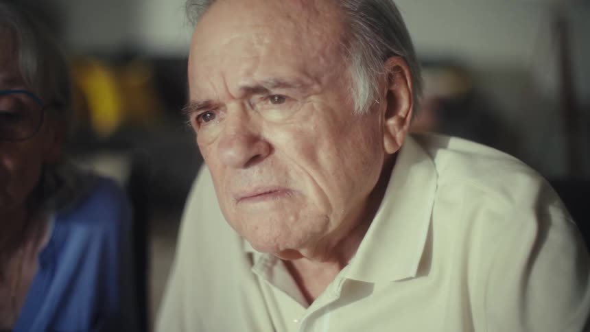 Elderly man in a light shirt looks intently at something in front of him with a serious expression