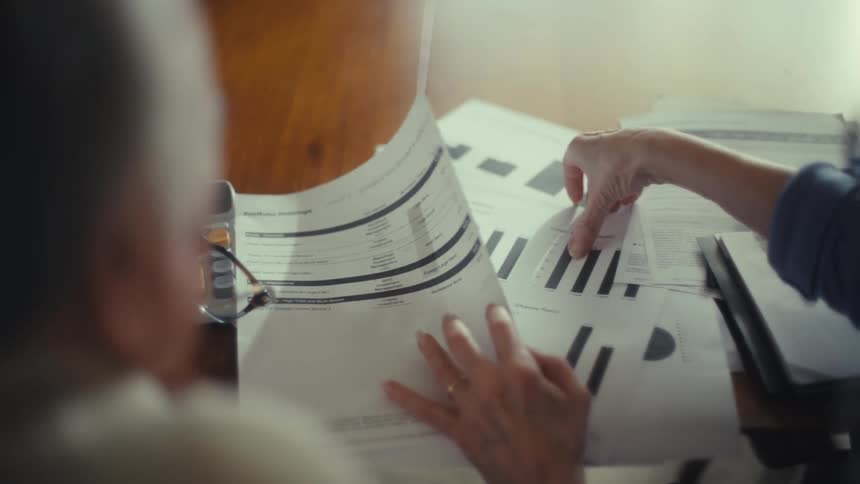 A man and a woman are seated at a table covered in papers as the woman holds up a document with graphs and text and points to a section with her finger