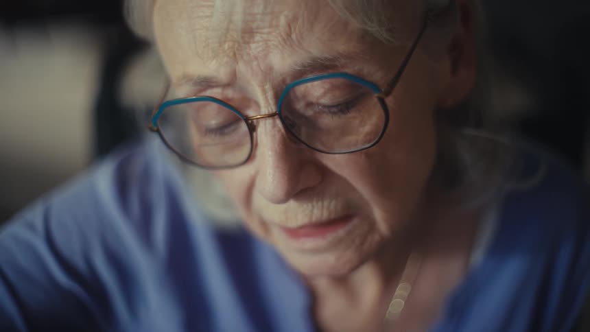 An older woman with gray hair and glasses is sitting at a table with papers and a calculator shes holding a pen and a small notebook looking intently at the papers on the table she has a concerned expression on her face
