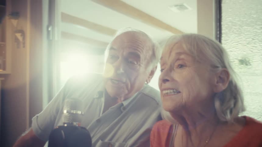 An elderly couple sits in their living room at midday with the woman in a wheelchair and the man beside her both singing karaoke into a microphone while laughing and smiling happily as sunlight streams through the window into the room