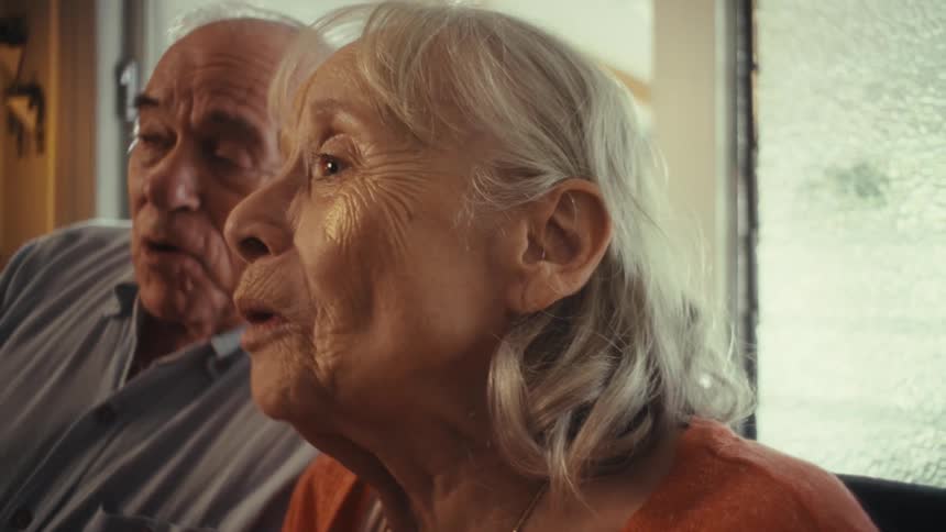 A smiling elderly woman with her mouth open and a man with a wide smile are both singing into a microphone as seen in a close view from the side