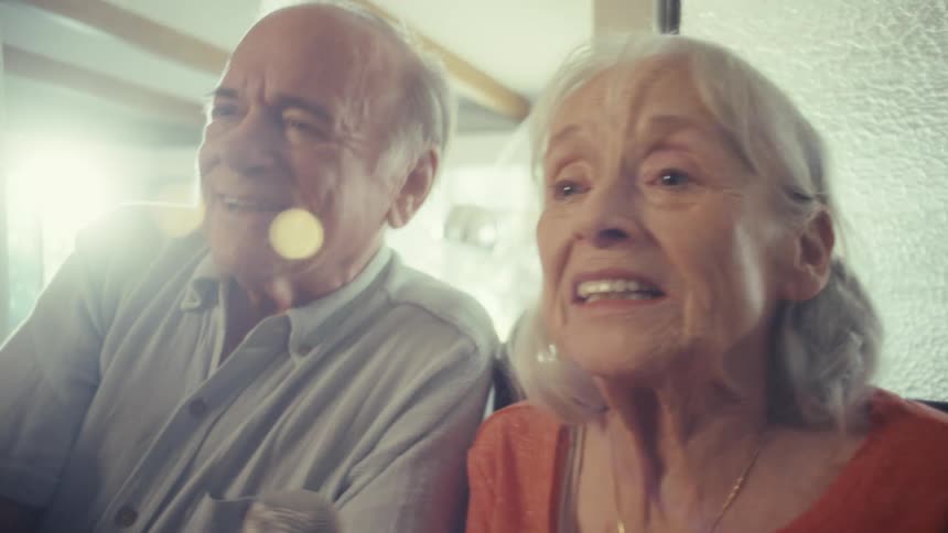 An older man and woman are sitting together the man is wearing a gray shirt and the woman is wearing an orange shirt with a necklace they are both looking to the side and the man has a serious expression while the woman appears to be speaking