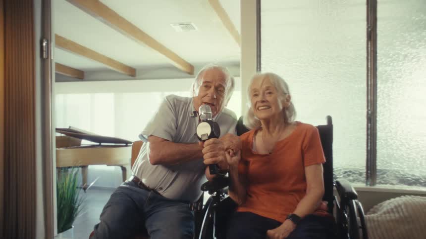 A man and woman sit next to each other in a room holding a microphone together looking at the camera and singing with faces that show happiness and enjoyment