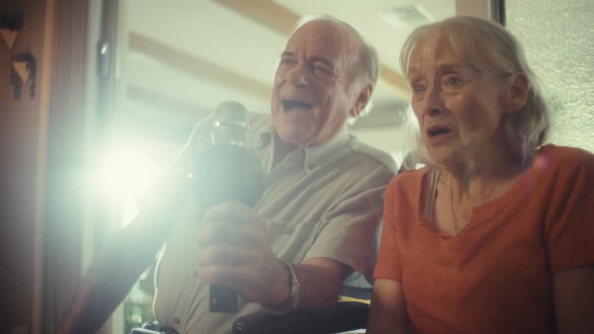 Elderly couple smiles and sings into a microphone while sitting at home during the daytime