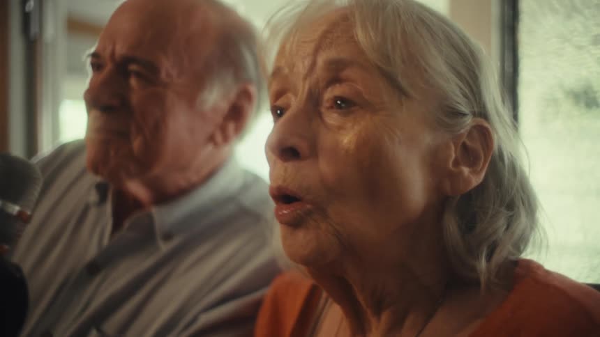 An elderly couple shares a microphone singing and having a good time in their living room at midday