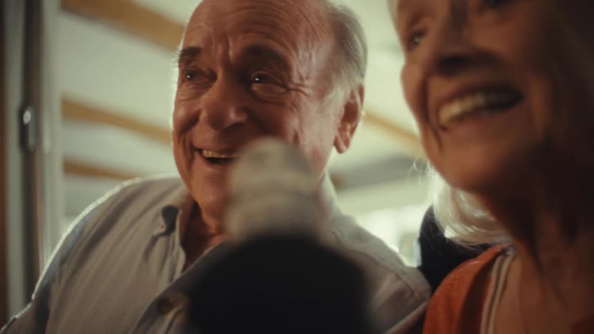 A close view shows a smiling elderly woman and a man both singing into a microphone with a bright light shining on their faces from the left