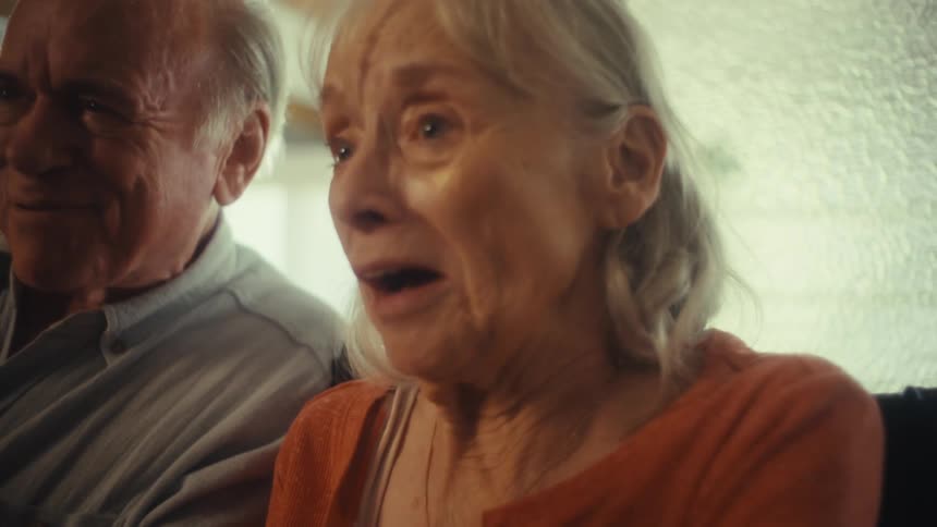 An elderly couple sits in their living room at midday with the woman in a wheelchair and the man beside her both singing karaoke into a microphone while laughing and smiling happily as sunlight streams through the window into the room