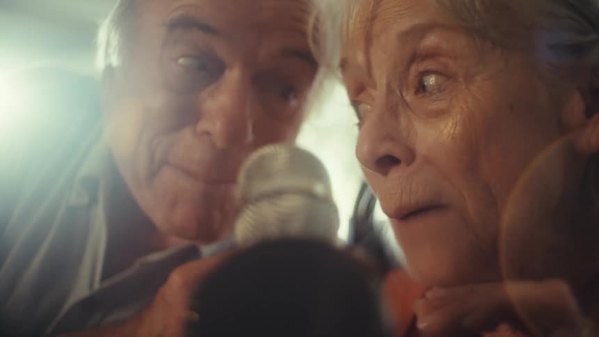 An older couple with gray hair is sitting close together and laughing the woman is in a wheelchair and is wearing an orange shirt while the man is wearing a gray shirt a bright light is shining from the side and the man is holding a microphone