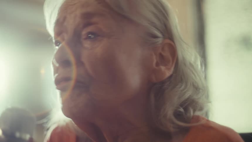 A closeup shot shows an elderly woman with white hair singing into a microphone she smiles broadly as she sings and a bright light from a window behind her shines on the scene