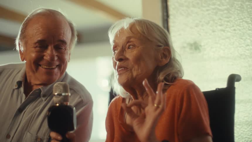 A woman with gray hair is seated in a wheelchair next to a man with a microphone and they are both smiling and looking to the left of the camera
