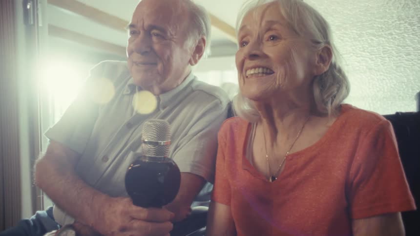 An older couple is sitting sidebyside holding a microphone the woman is in a wheelchair and is wearing an orange shirt while the man is sitting on a couch they are singing and laughing happily together