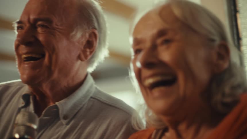 A close view of a smiling elderly man and woman shows them both singing into a microphone with happy expressions on their faces