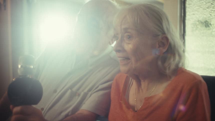 An elderly couple joyfully sings karaoke together into a microphone that flashes with a bright light in their living room