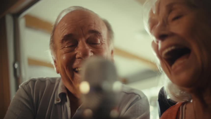 An elderly couple sits side by side in the living room at midday happily singing karaoke into a microphone