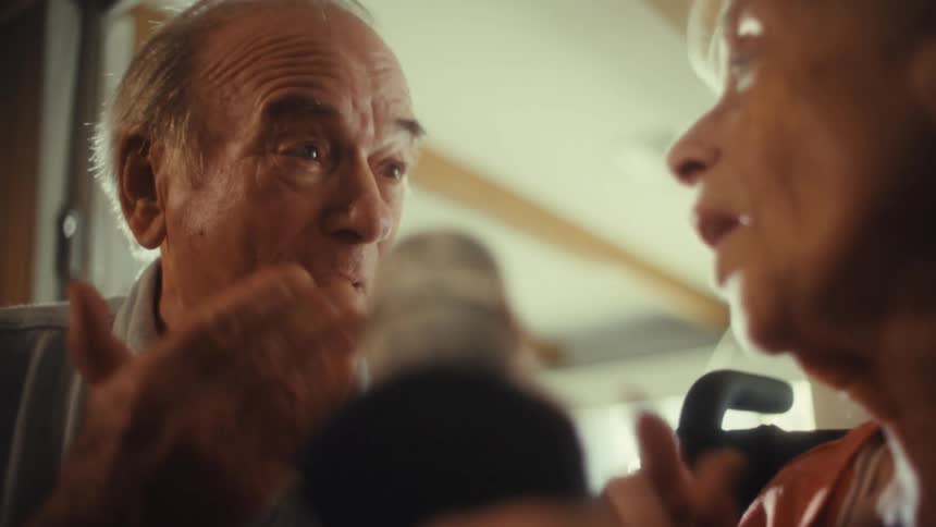An elderly couple sits in their living room at midday with the woman in a wheelchair and the man beside her both singing into a microphone happily and excitedly